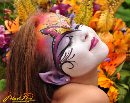 long haired girl in front of colorful flowers with a mask painted on her face in bright yellow pink and purple with elf like ears